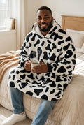Man wearing a CozyNest black and white cow print plush fleece wearable blanket hoodie with a sherpa-lined hood.