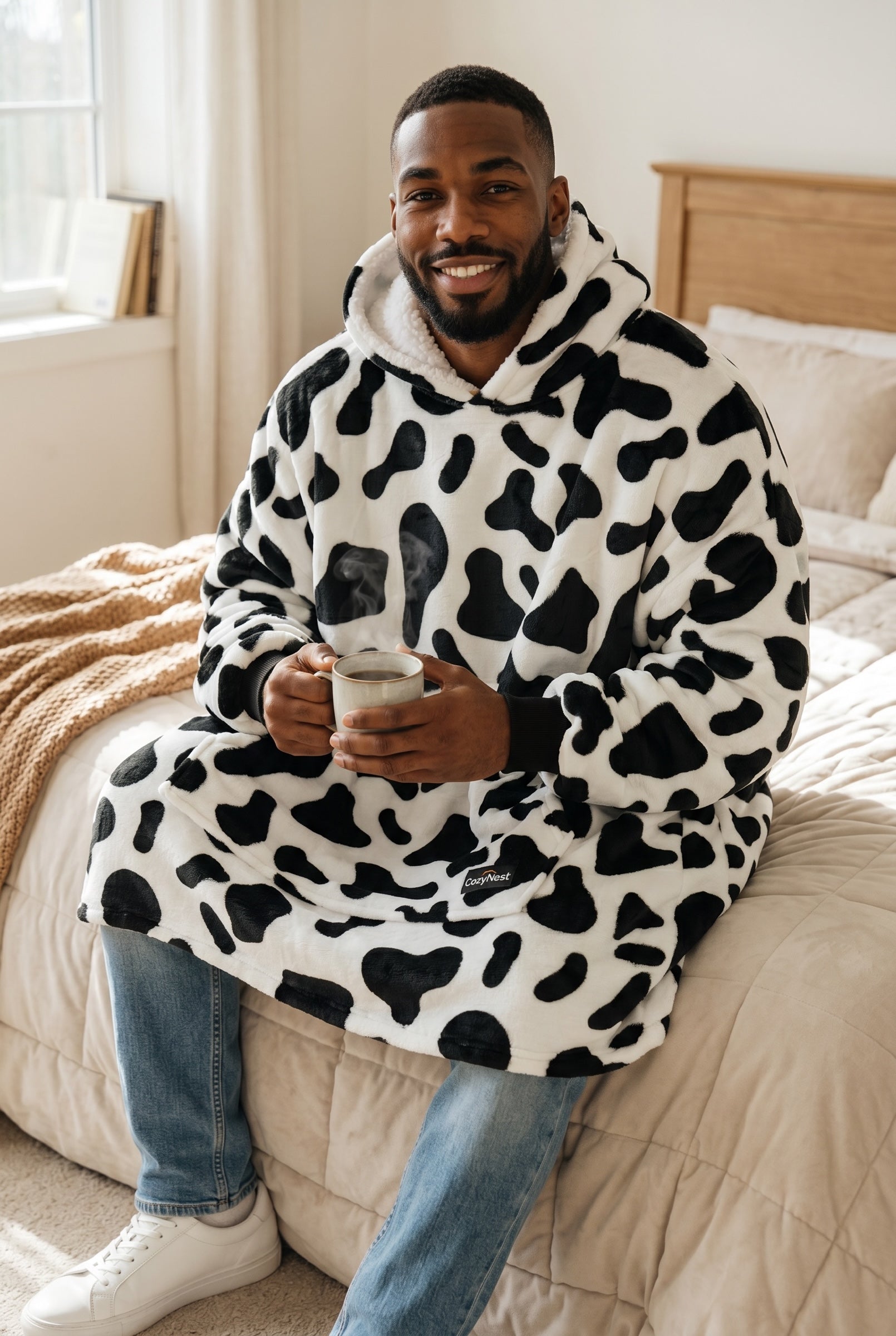 Man wearing a CozyNest black and white cow print plush fleece wearable blanket hoodie with a sherpa-lined hood.